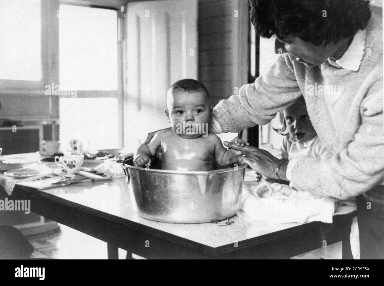 A young Australian mother bathes her new baby boy in a large jam pot on a kitchen table while his older sister watches on with intense interest Stock Photo