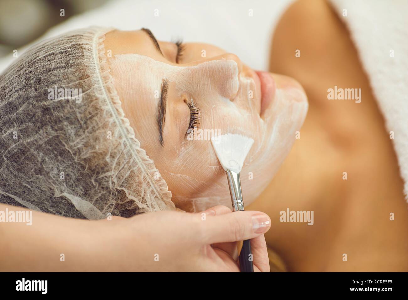 Young serene woman getting skincare procedure of applying moisturizing mask with brush from therapist Stock Photo