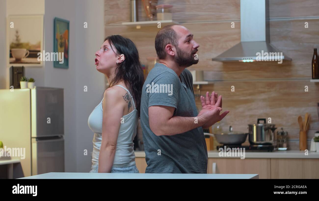 Couple with trust issues fighting back to back. Furious, irritated, frustrated, jealous unhappy couple screaming accusing to each other having family conflict disputing sitting in the kitchen. Stock Photo