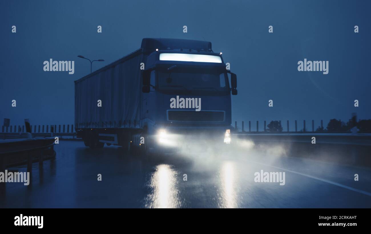 Blue Long Haul Semi-Truck with Cargo Trailer Full of Goods Travels At Night on the Freeway Road, Driving Across Continent Through Rain, Fog, Snow Stock Photo
