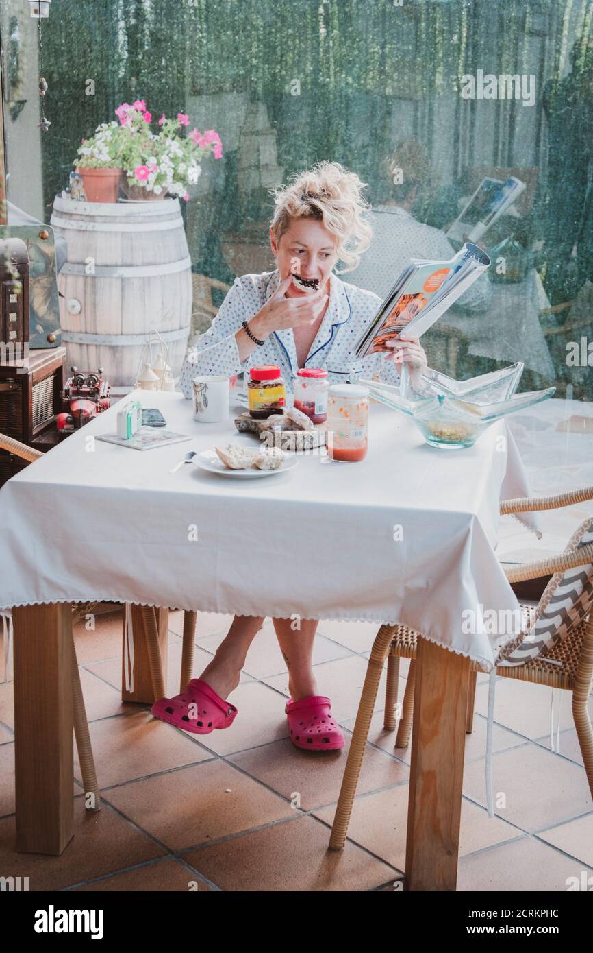 Blonde young mature woman in pyjamas at home in breakfast time, reading a magazine and eating a toast. Stock Photo