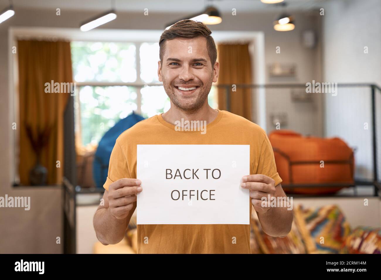 Reopening Business after lockdown. Young happy man showing paper with text BACK TO OFFICE at camera and smiling while standing in the modern coworking Stock Photo