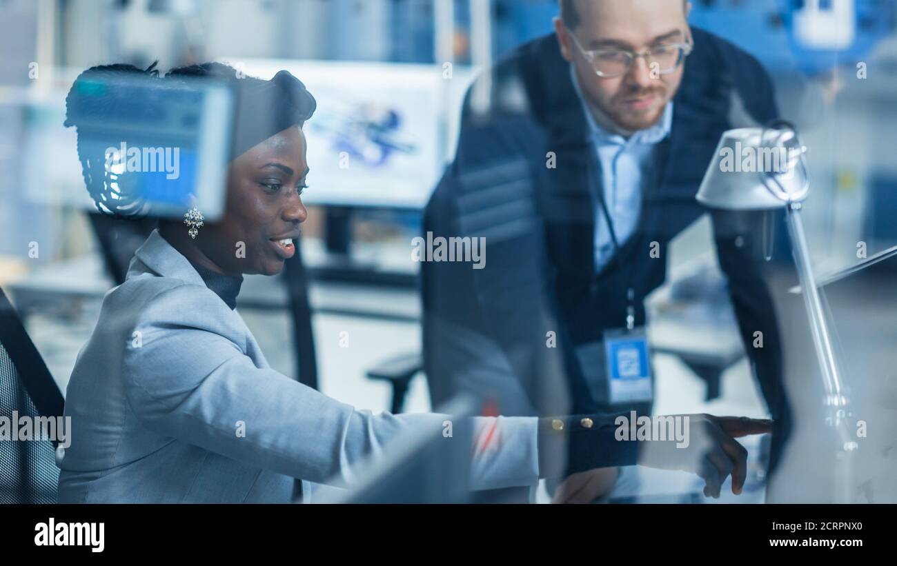 Shot Through Glass in Modern Electronics Factory: Female Supervisor Talks to a Male Electrical Engineer who Works on Computer. Designing PCB Stock Photo