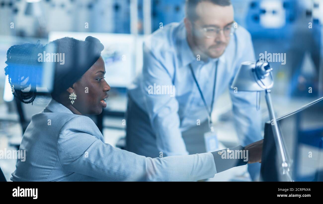 Modern Factory: Male Project Supervisor Talks to a Female Industrial Engineer who Works on Personal Computer. Shot Through Glass in Modern High-Tech Stock Photo
