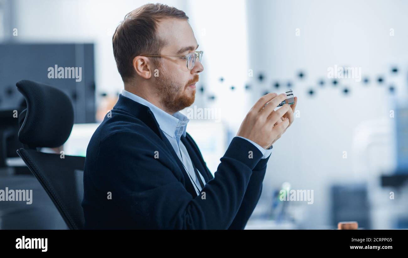 Electronics Factory: Male Electrical Engineer Holds PCB Prototype, Works on Personal Computer. Modern High-Tech Industrial Factory Office. Stock Photo