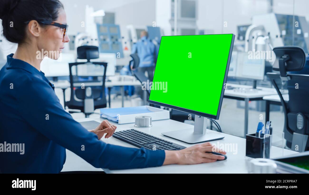 Over the Shoulder: Female Chief Engineer Working on Personal Computer, Display is Green Mock-up Screen or Chroma Key. Industrial Factory with High Stock Photo
