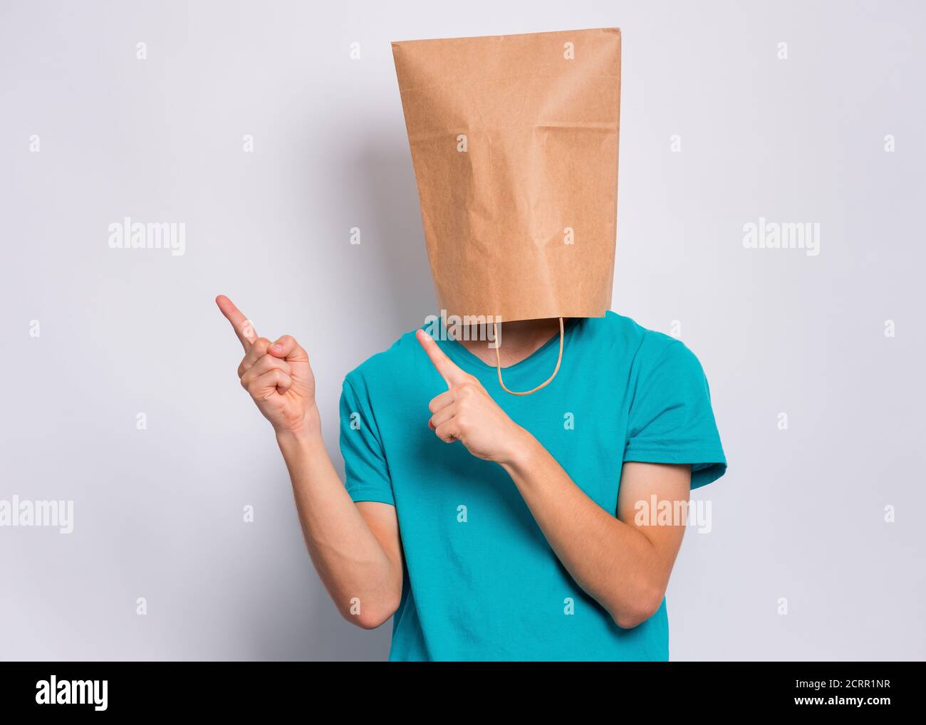 Teen boy with paper bag over head pointing finger away at copyspace, on gray background. Teenager cover head with shopping bag pointing finger at some Stock Photo