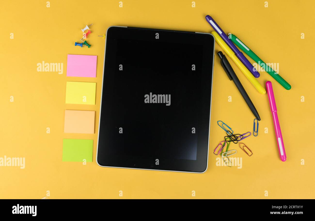 Top view of a tablet and school supplies like colored markers, sticker and clipers on yellow background, space for text. Stock Photo