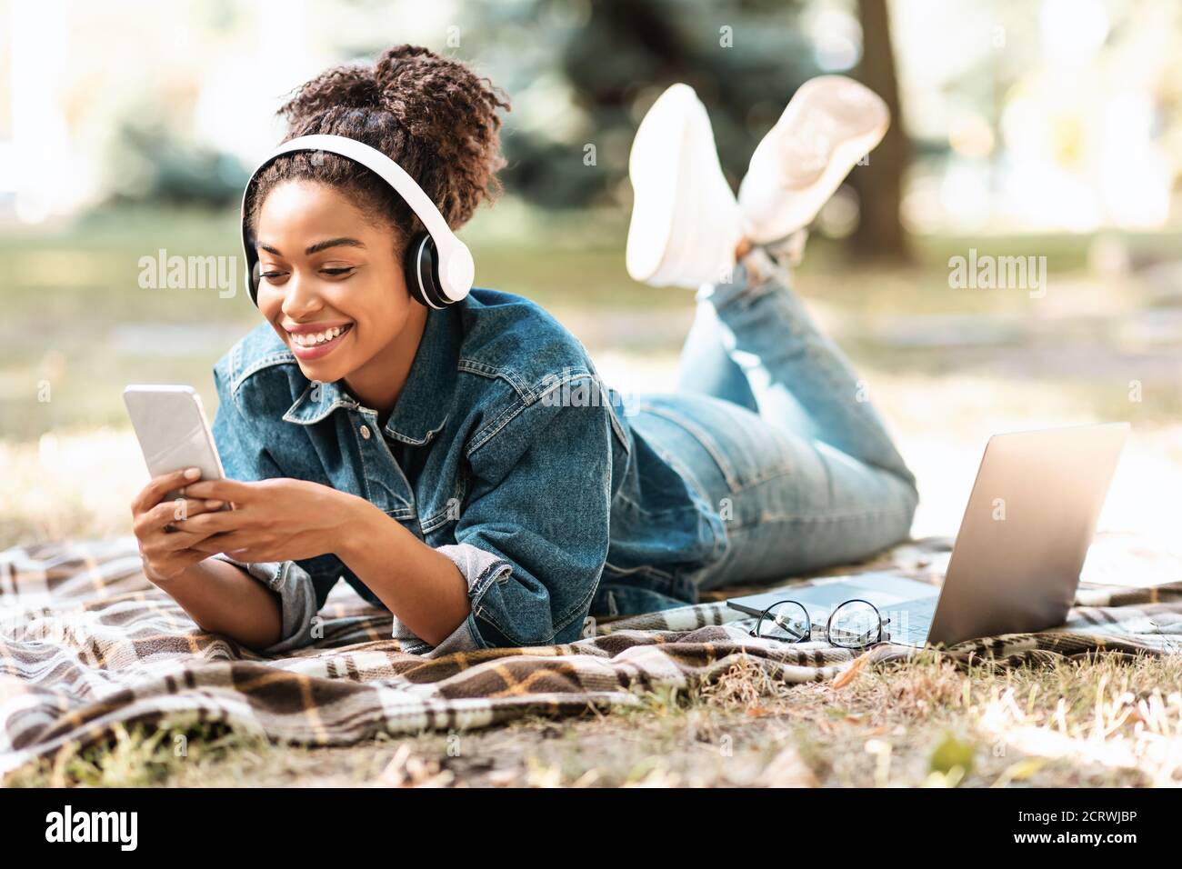Cheerful Black Lady Using Phone Learning Lying On Plaid Outdoors Stock Photo