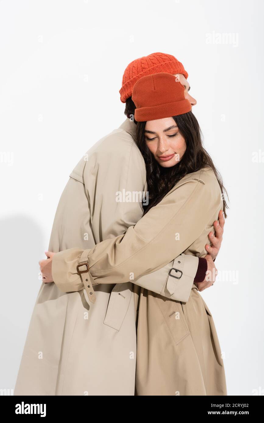 stylish couple in beanie hats and trench coats hugging on white Stock ...