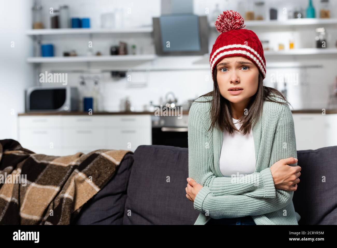 freezing woman in knitted hat hugging herself while sitting in cold kitchen Stock Photo