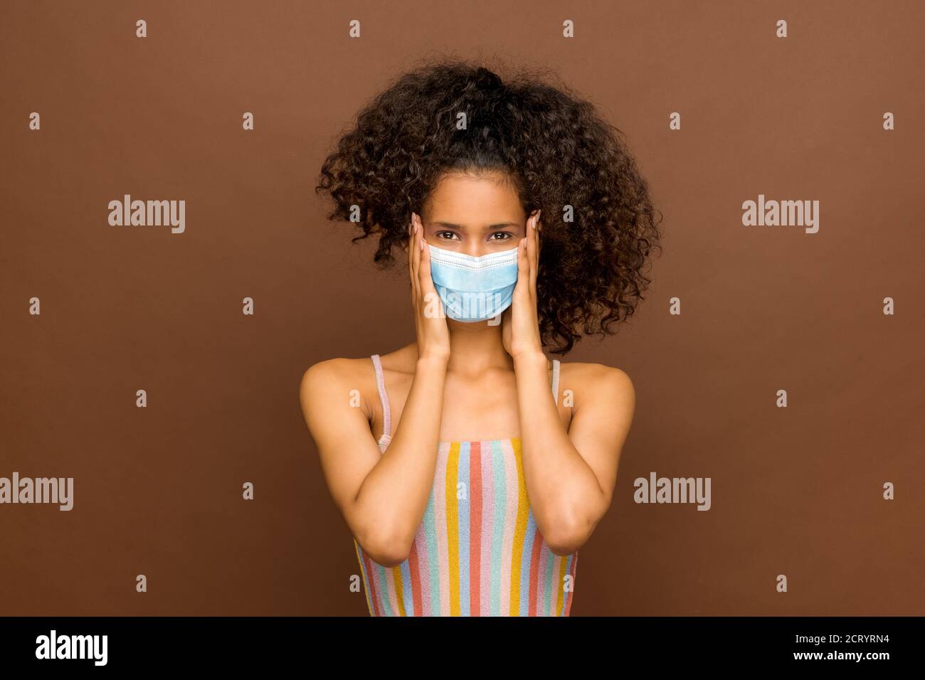 Attractive Dominican girl with thick curly long hair wearing a protective face mask for infection control during the Covid-19 pandemic over a brown st Stock Photo