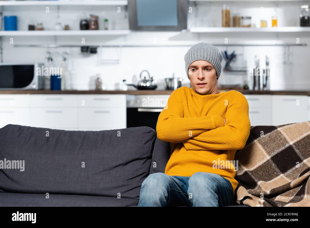 shivering man in knitted sweater and hat sitting on sofa with crossed arms in cold kitchen Stock Photo