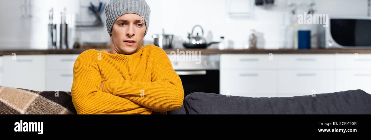 horizontal crop of freezing man in warm sweater and hat sitting with crossed arms in cold kitchen Stock Photo