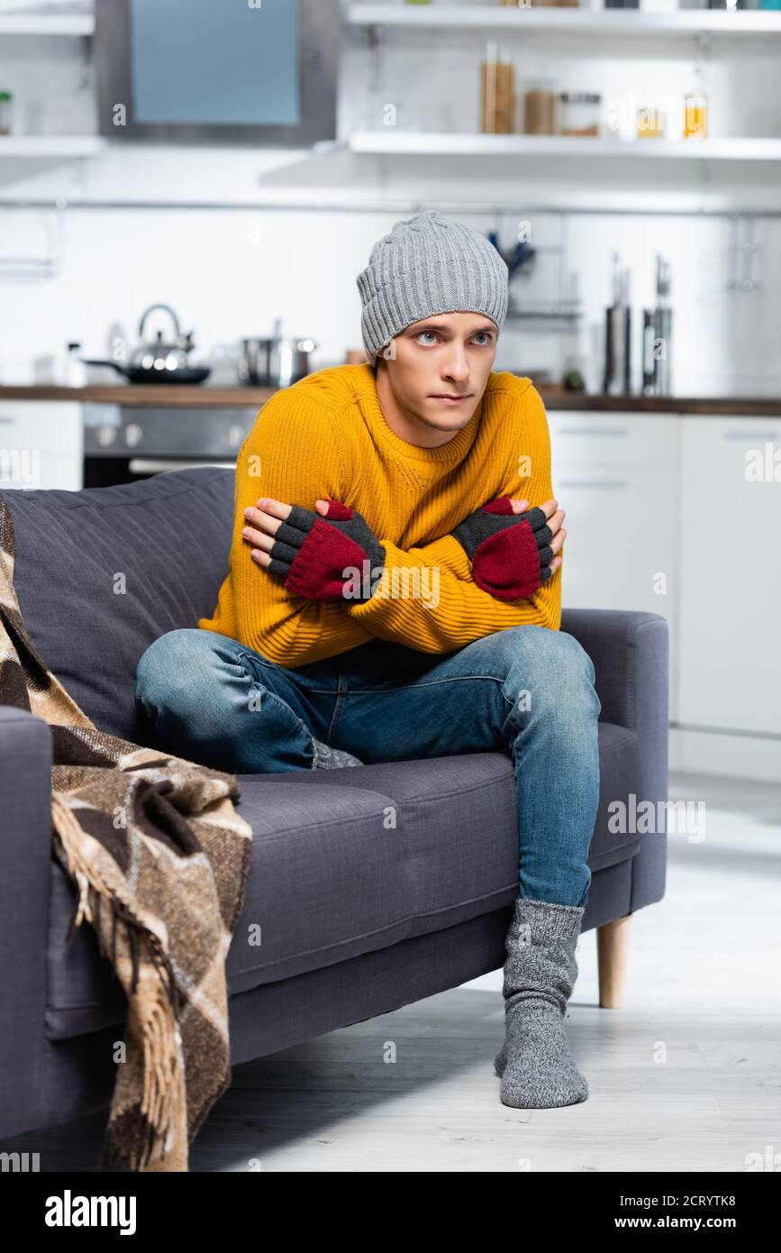 freezing man in warm hat and fingerless gloves hugging himself while sitting on sofa in cold kitchen Stock Photo