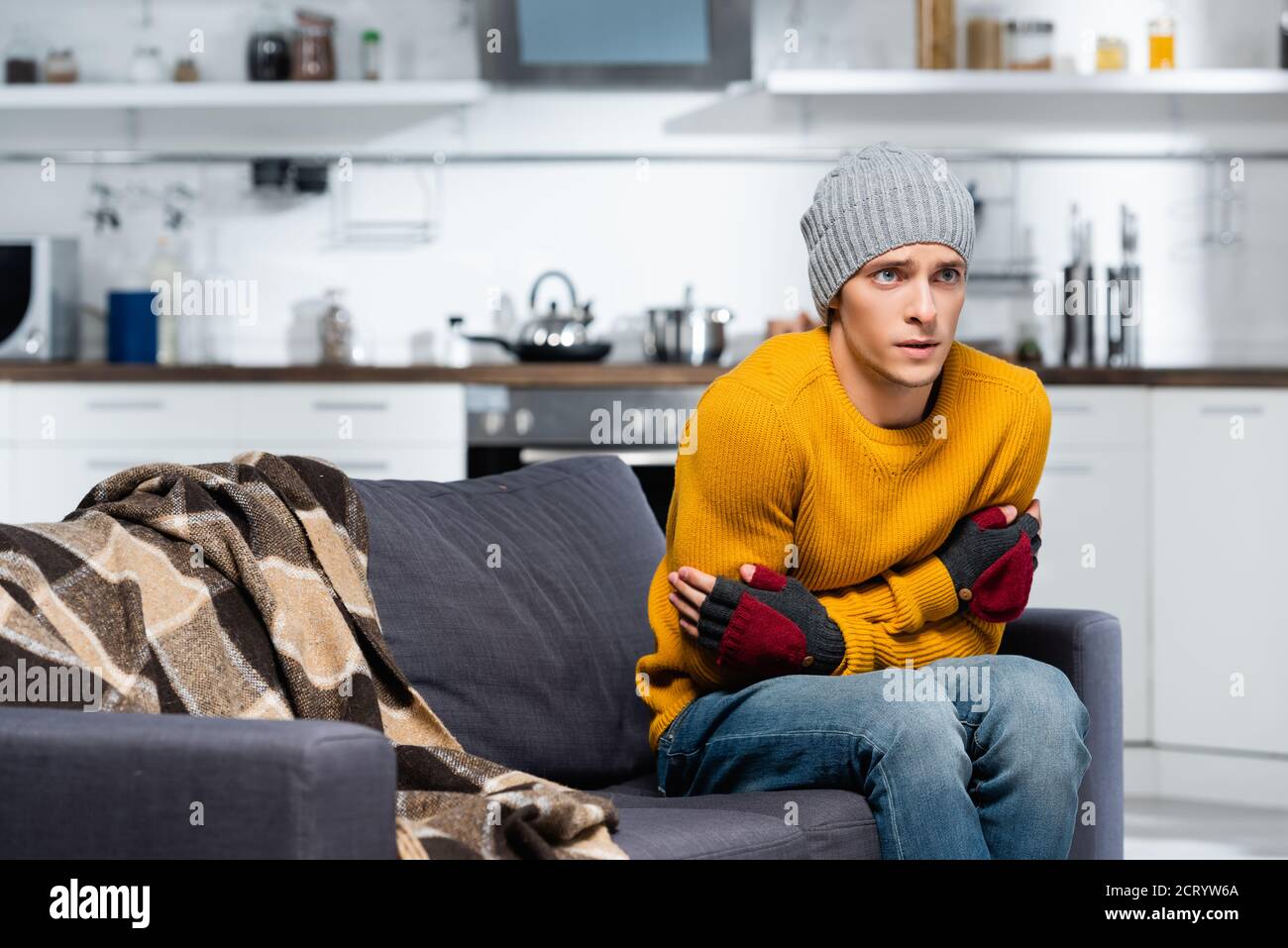 young man in warm hat and fingerless gloves hugging himself while sitting on sofa in cold kitchen Stock Photo