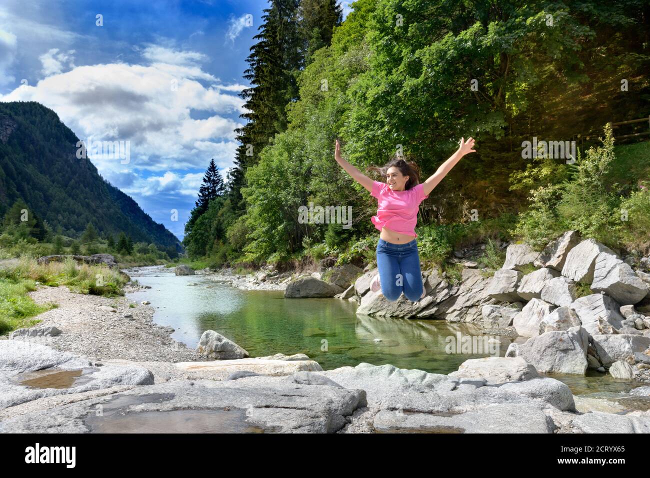 Energetic young girl celebrating her freedom jumping in the air with outstretched arms on rocks alongside a river in a mountain valley Stock Photo