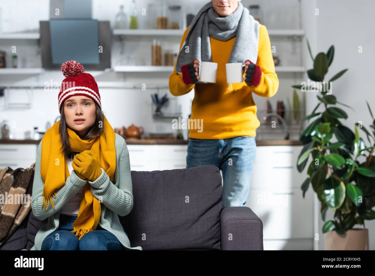 freezing woman sitting on sofa in knitted hat and gloves while man ...