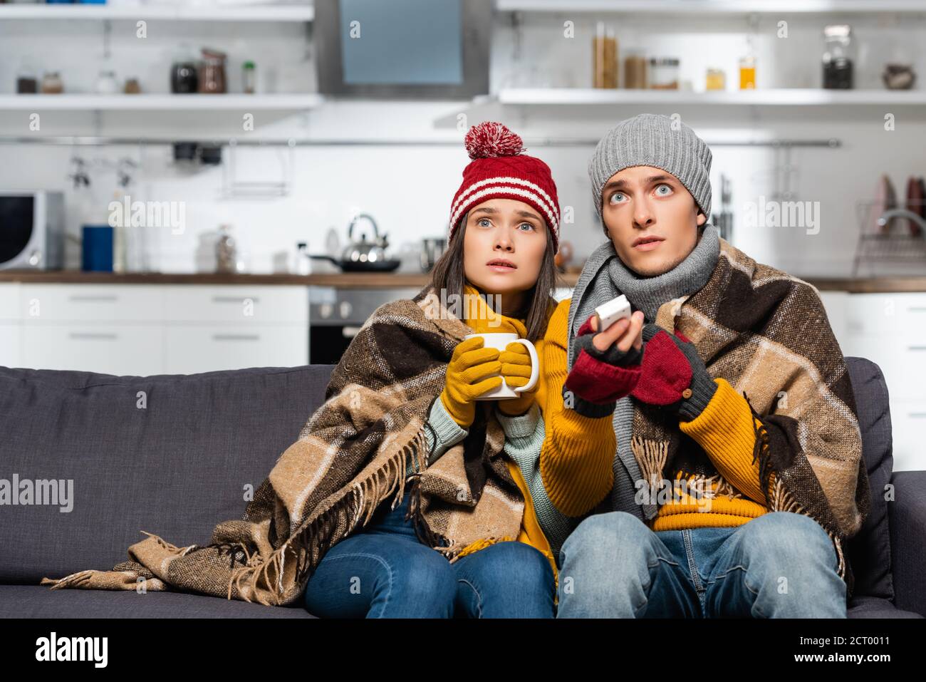 freezing-couple-in-warm-hats-wrapped-in-plaid-blanket-watching-tv-while-sitting-in-cold-kitchen-2CT0011.jpg