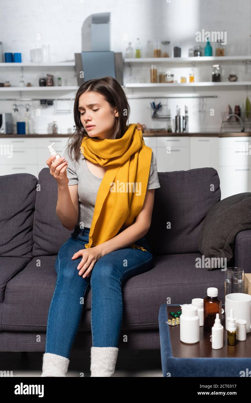 sick woman with scarf on neck holding throat spray while sitting near bedside table with medicines Stock Photo