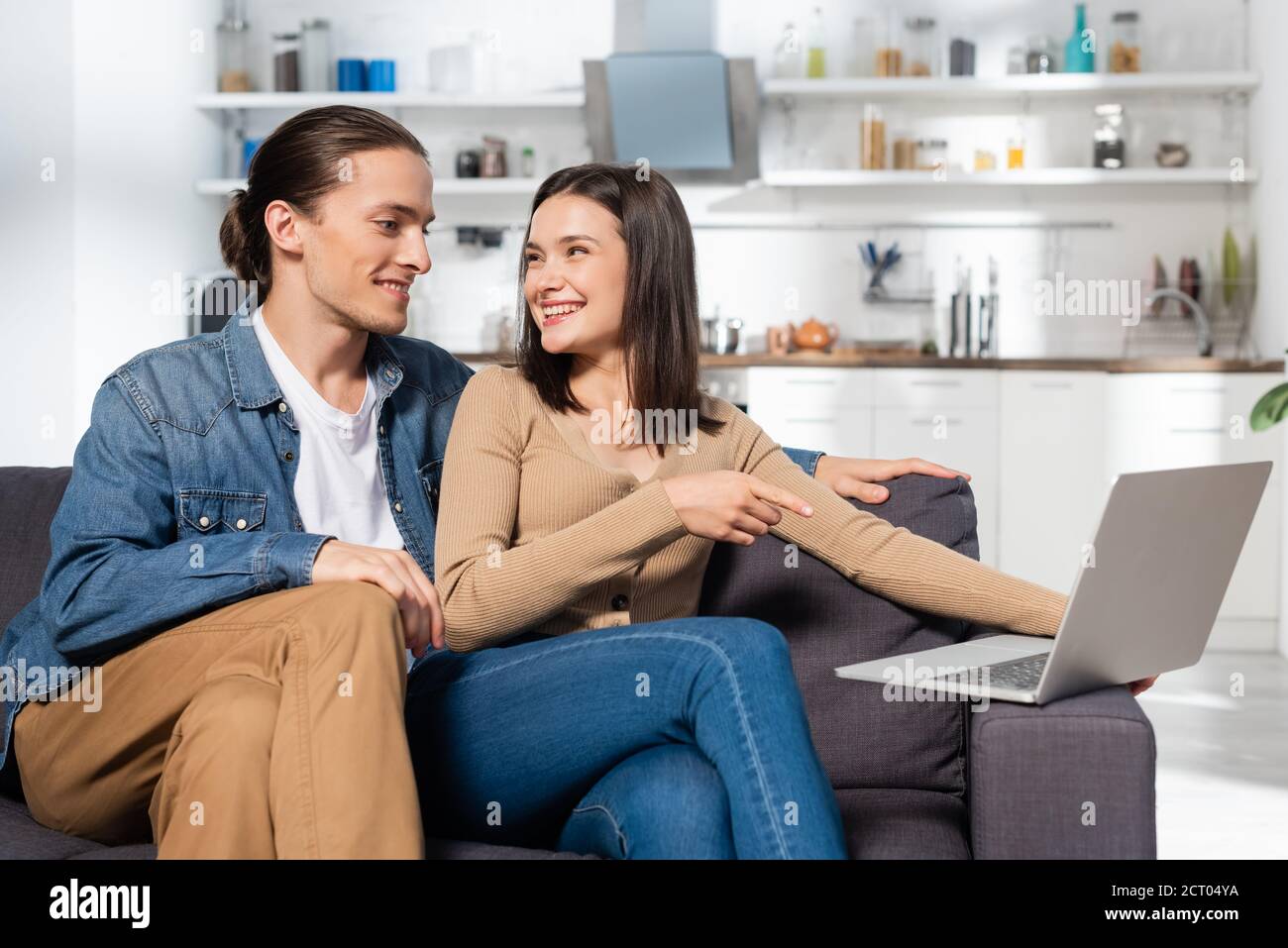 man sitting near excited girlfriend pointing with finger at laptop on couch in kitchen Stock Photo
