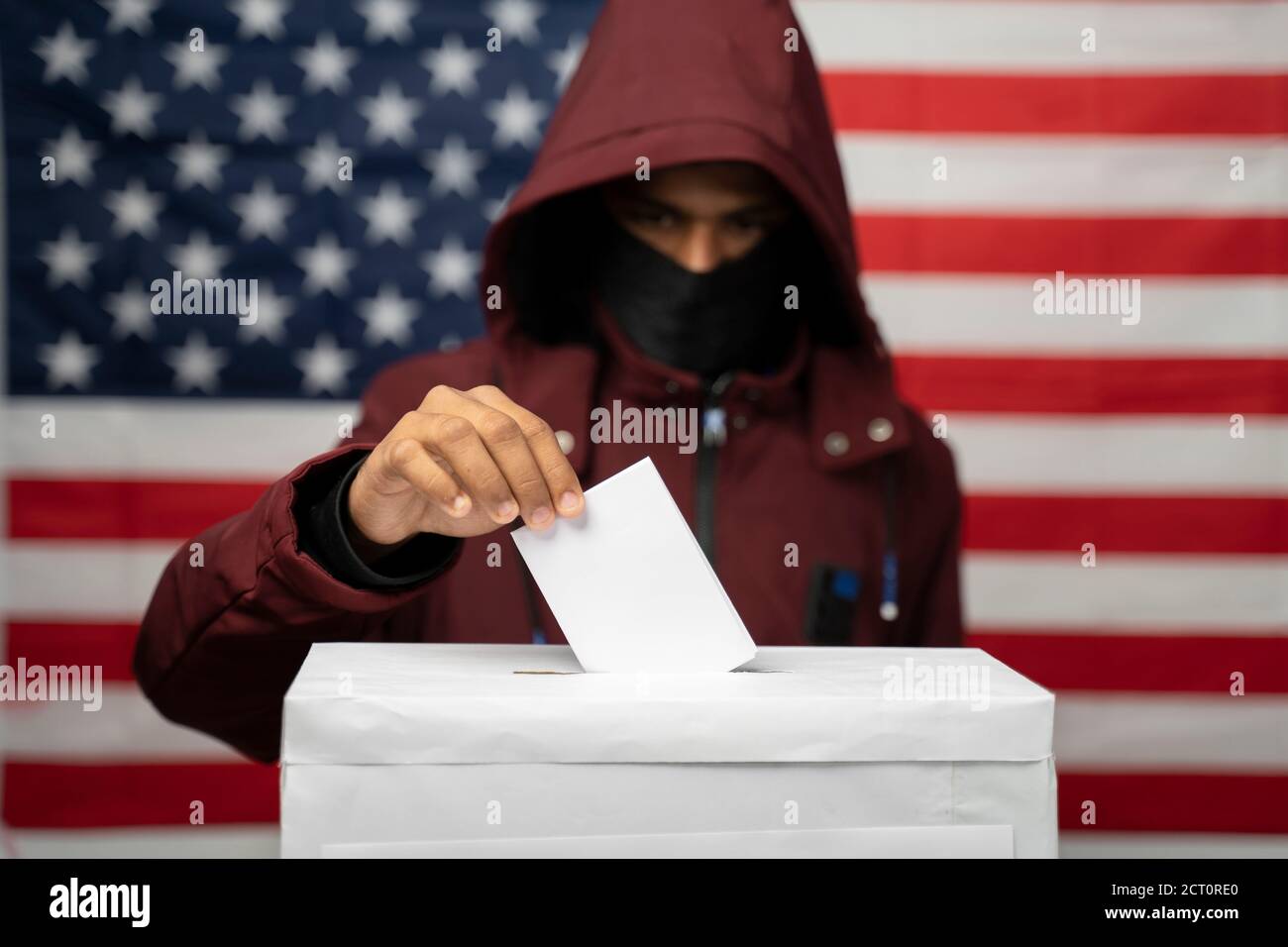 Man in hoodie with face covered casting Vote at polling booth with US falg as background - Concept of unkonwn voting or vote rigging in US elections Stock Photo