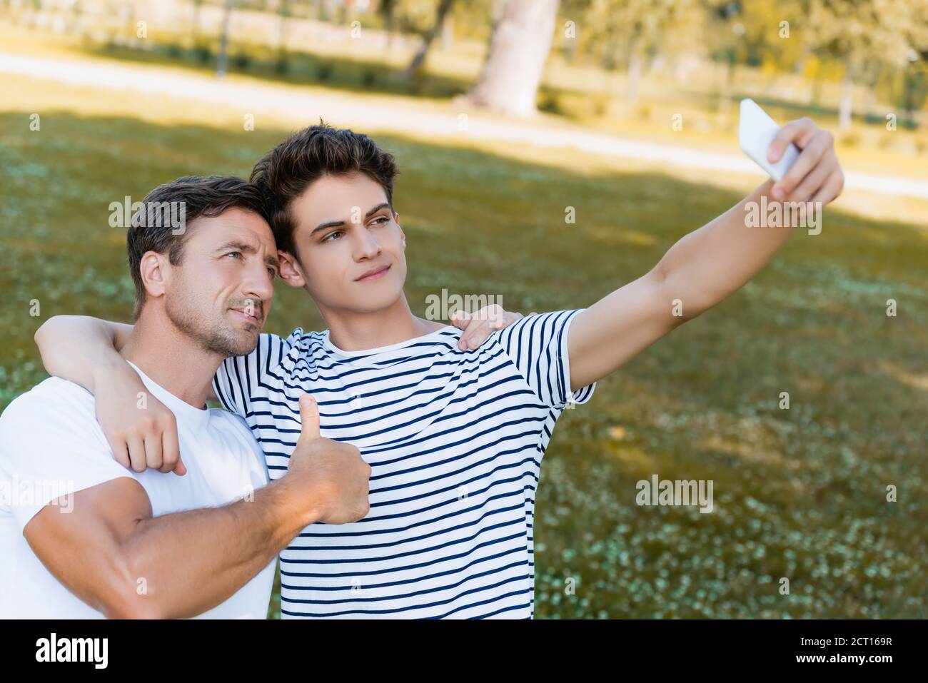father showing thumb up and teenager son taking selfie in park Stock Photo