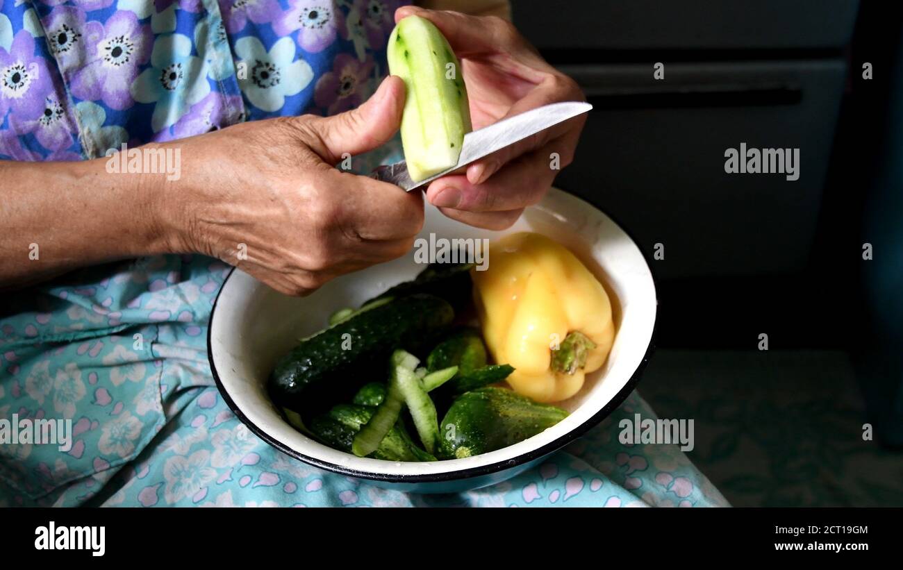 Granny hands cut green cucumber with knife. 80 year old woman hands prepare veggie food in rustic kitchen Stock Photo