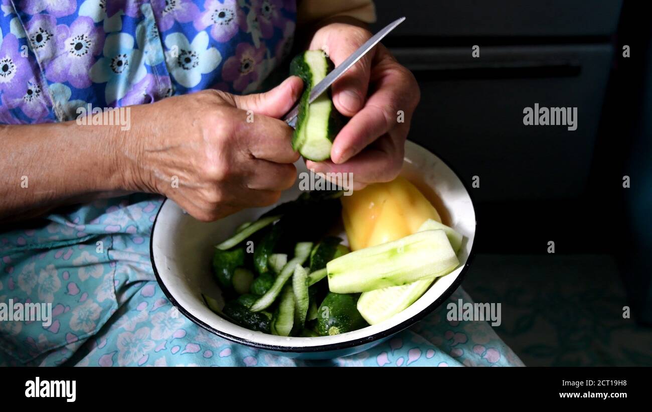 Granny hands peel fresh cucumber with knife and cut into pieces. Authentic scene of 80 year old woman preparing food in rustic kitchen Stock Photo