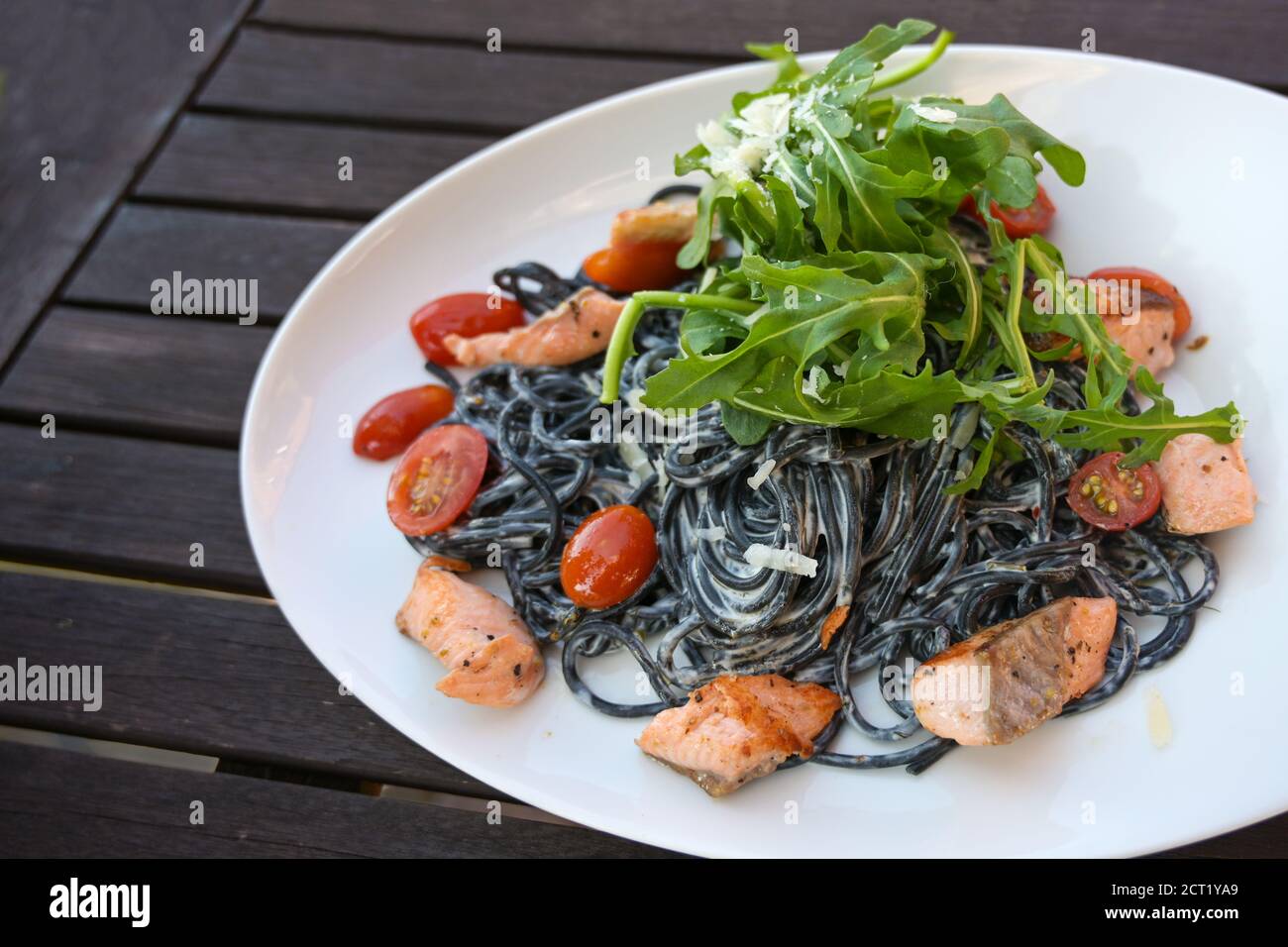 Black spaghetti pasta colored with squid ink, cream sauce, salmon, tomatoes and rocket salad in a white plate on a dark rustic wooden garden table, se Stock Photo