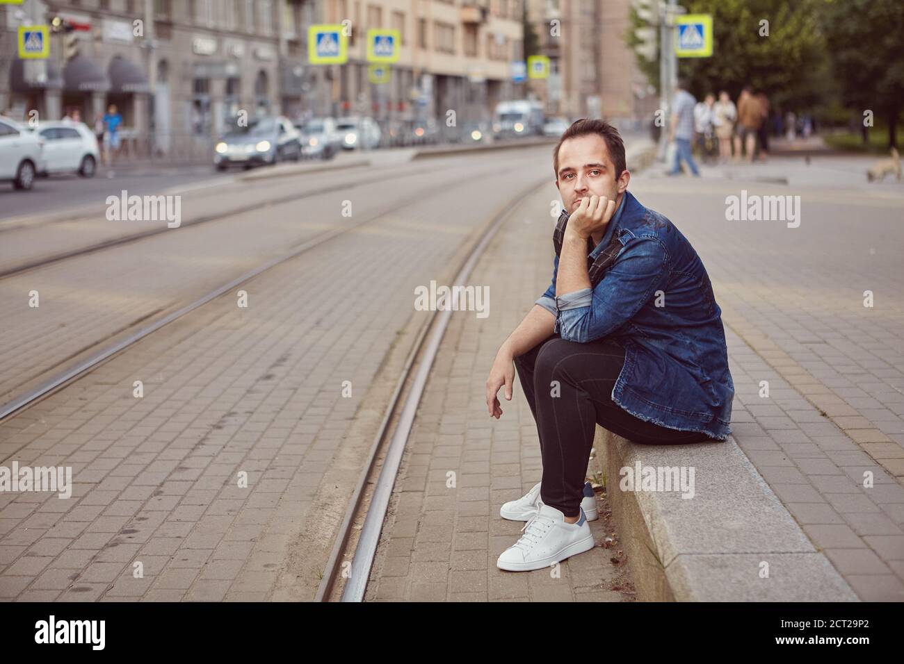 A man is sitting on the sidewalk near the railroad. Young white upset male in the city. Stock Photo
