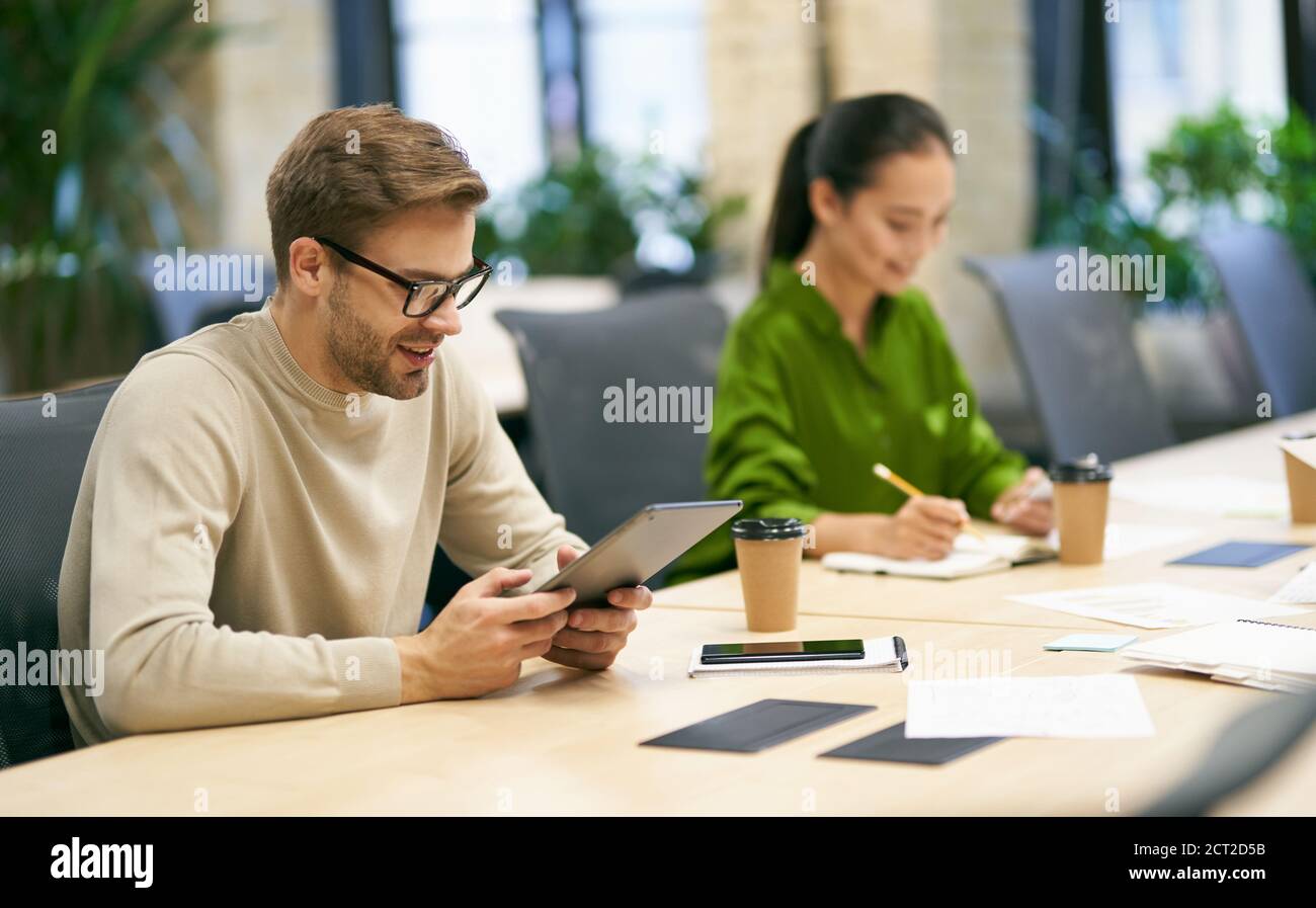 Working together. Young smiling caucasian man wearing eyeglasses using digital tablet while sitting at desk with his female colleague in the modern coworking space. Business people and teamwork Stock Photo