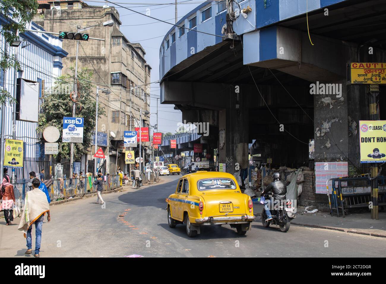 Kolkata, India - February 1, 2020: Unidentified people walks on the street as a traditional yellow taxi and motorbike drives by underneath Maa Flyover Stock Photo
