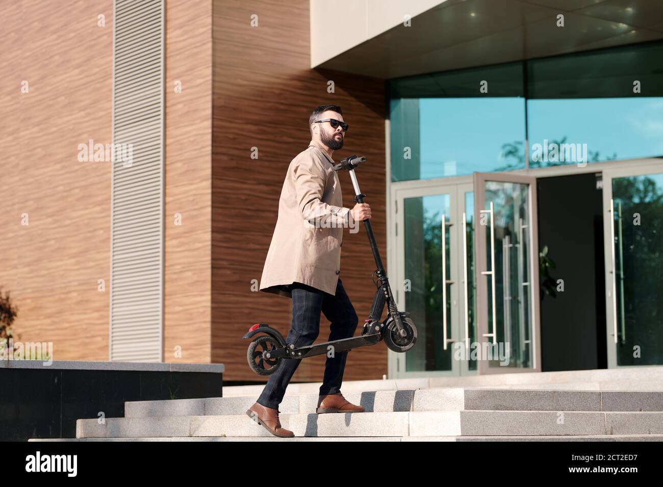 Young businessman in trenchcoat, pants and sunglasses carrying electric scooter Stock Photo