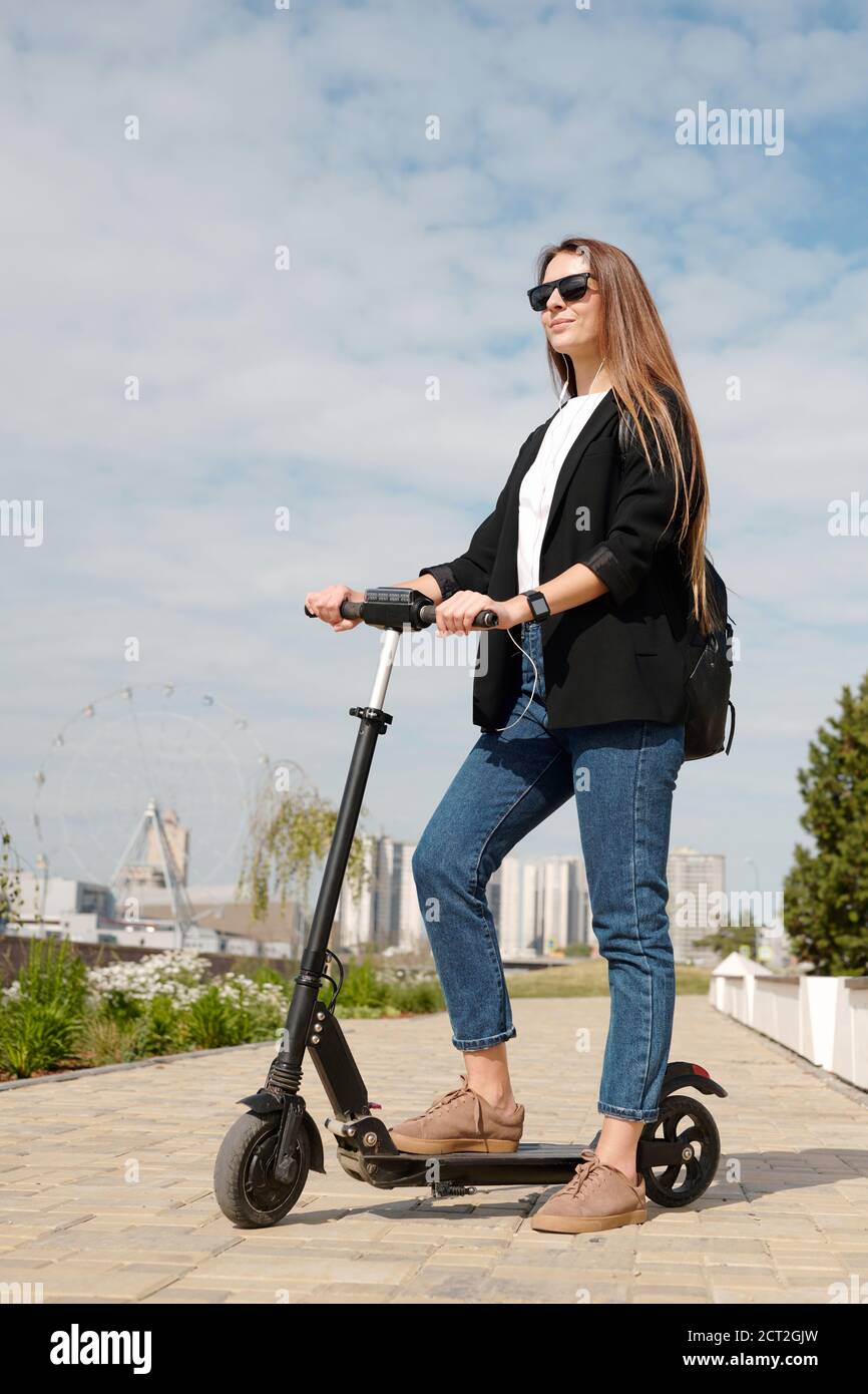 Elegant young businesswoman in casualwear standing on electric scooter on road Stock Photo