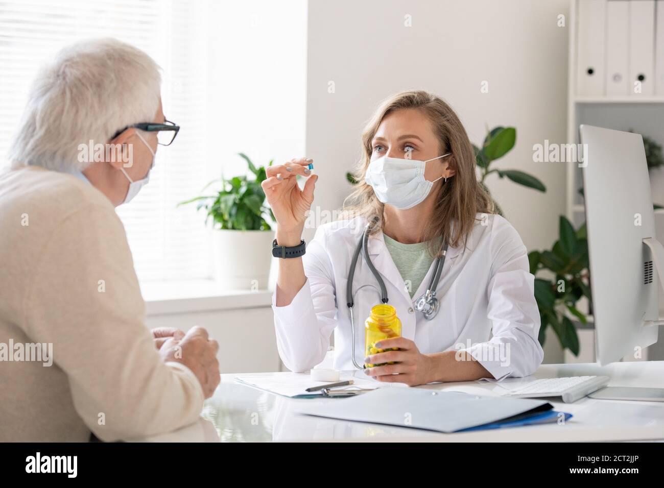 Young confident doctor in whitecoat and protective mask showing pill to patient Stock Photo