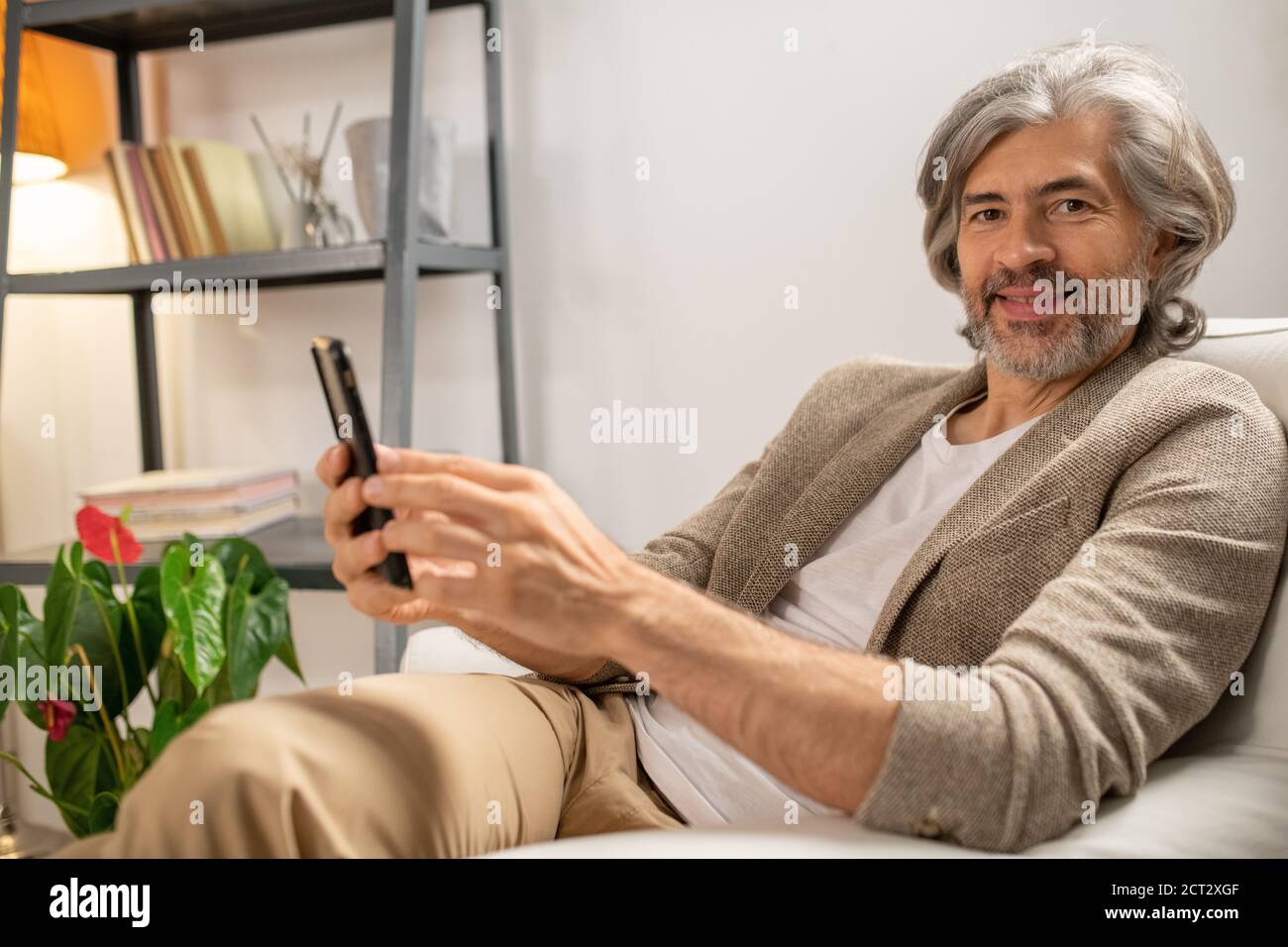 Happy mature bearded man looking at you with smile while sitting in armchair Stock Photo