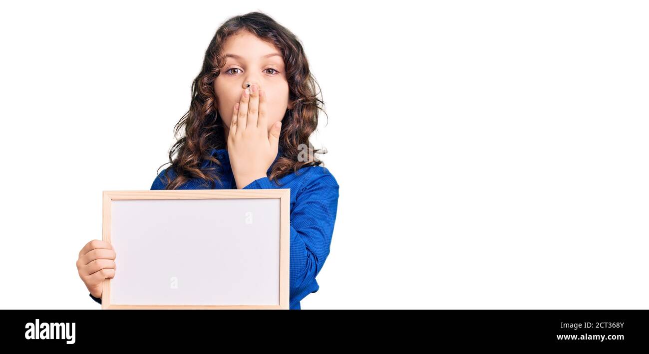 Cute hispanic child with long hair holding empty white chalkboard covering mouth with hand, shocked and afraid for mistake. surprised expression Stock Photo
