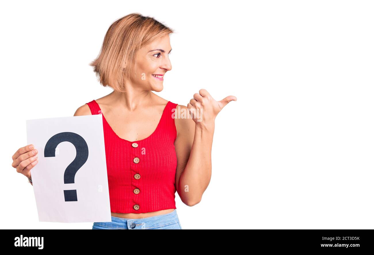 Young blonde woman holding question mark pointing thumb up to the side ...