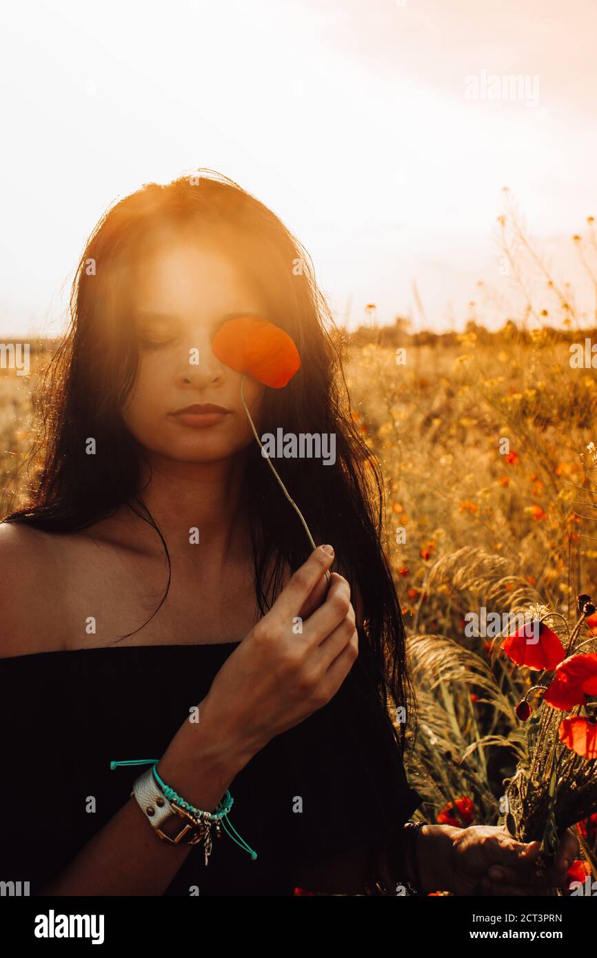 Summer portrait of beautiful tanned, brunette Ukrainian woman with long dark hair closing one eye with blooming red poppy and bouquet in hand in off s Stock Photo