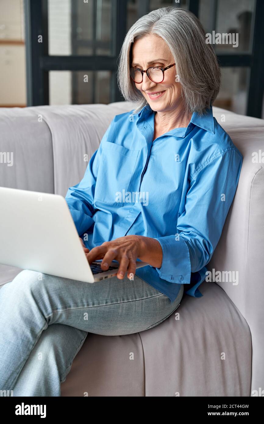 Happy 60s mature woman using laptop computer sitting on couch at home. Stock Photo