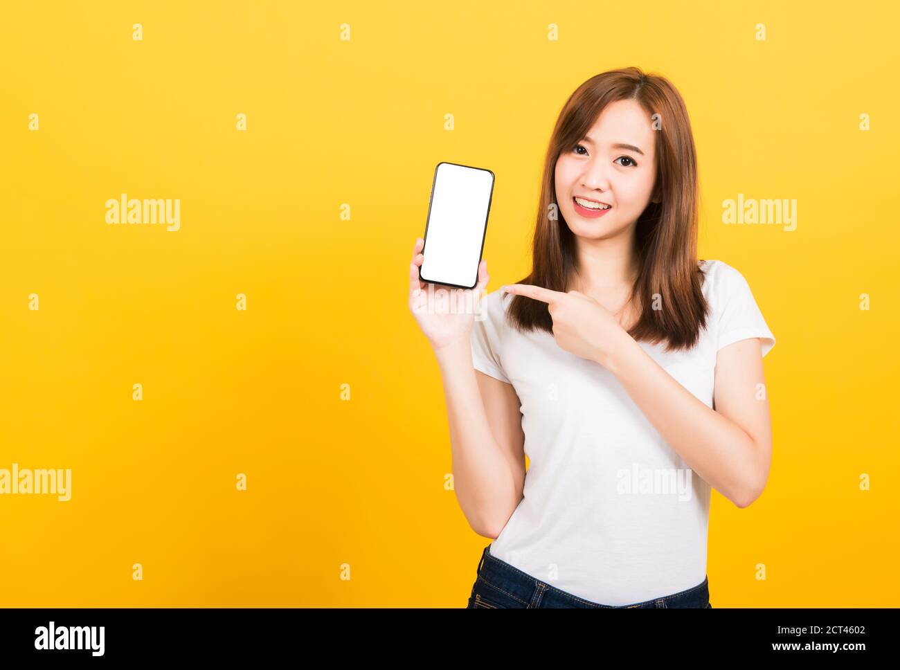 Asian happy portrait beautiful cute young woman smile standing wear t-shirt making finger pointing on smartphone blank screen looking to camera isolat Stock Photo