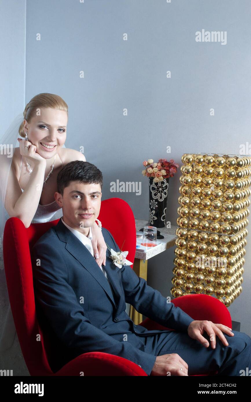 Groom sitting in red chair and smiling bride behind with empty wall for copy space Stock Photo