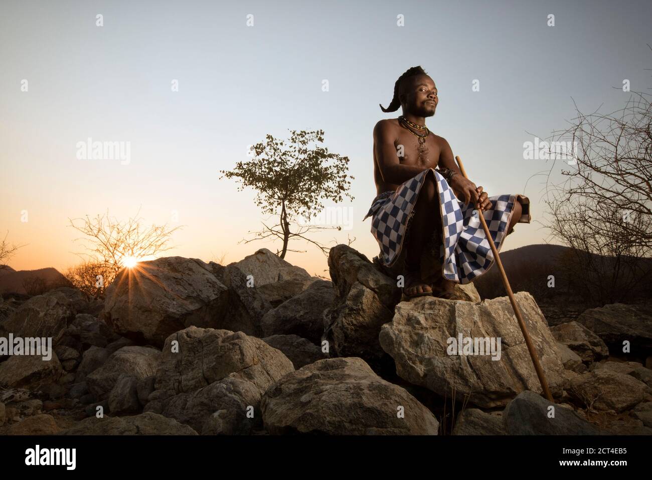 A Himba man sits on a rock as the sun sets at the end of the day in Epupa, Kunene Region, Namibia. Stock Photo