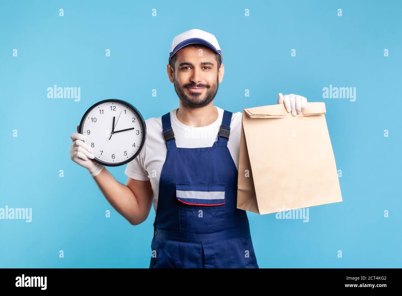 Food delivery, shipping on time! Portrait of handyman in uniform, gloves holding clock and parcel. Express courier transportation, post mail services. Stock Photo