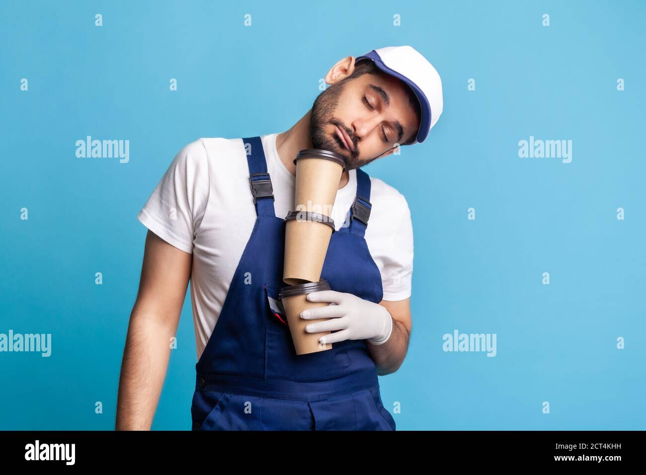 Delivery service. Tired inefficient courier man in overalls lying on coffee cups and sleeping, feeling exhausted bored after stressful night shift. La Stock Photo