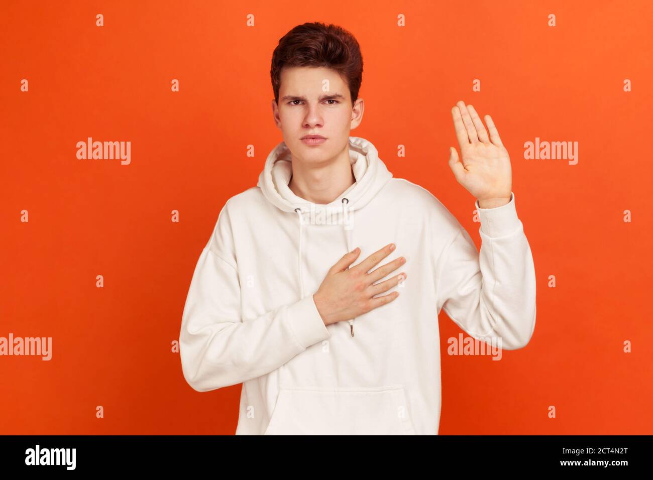 Serious patriotic male in white sweatshirt with hood holding hand on heart, juryman swearing to speak truth in court, honor and conscience. Indoor stu Stock Photo