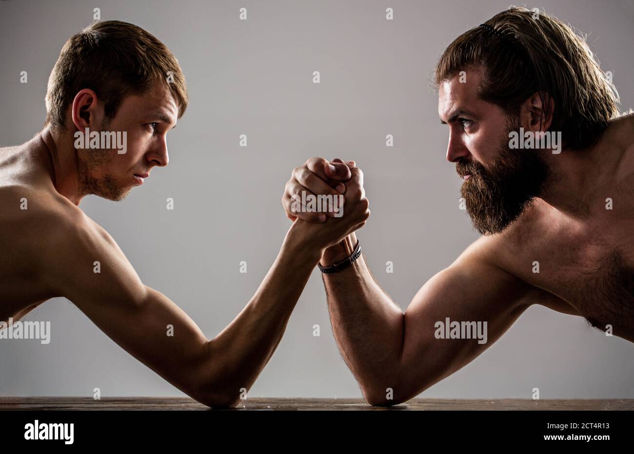 Arms wrestling thin hand, big strong arm in studio. Two man's hands clasped arm wrestling, strong and weak, unequal match. Heavily muscled bearded man Stock Photo