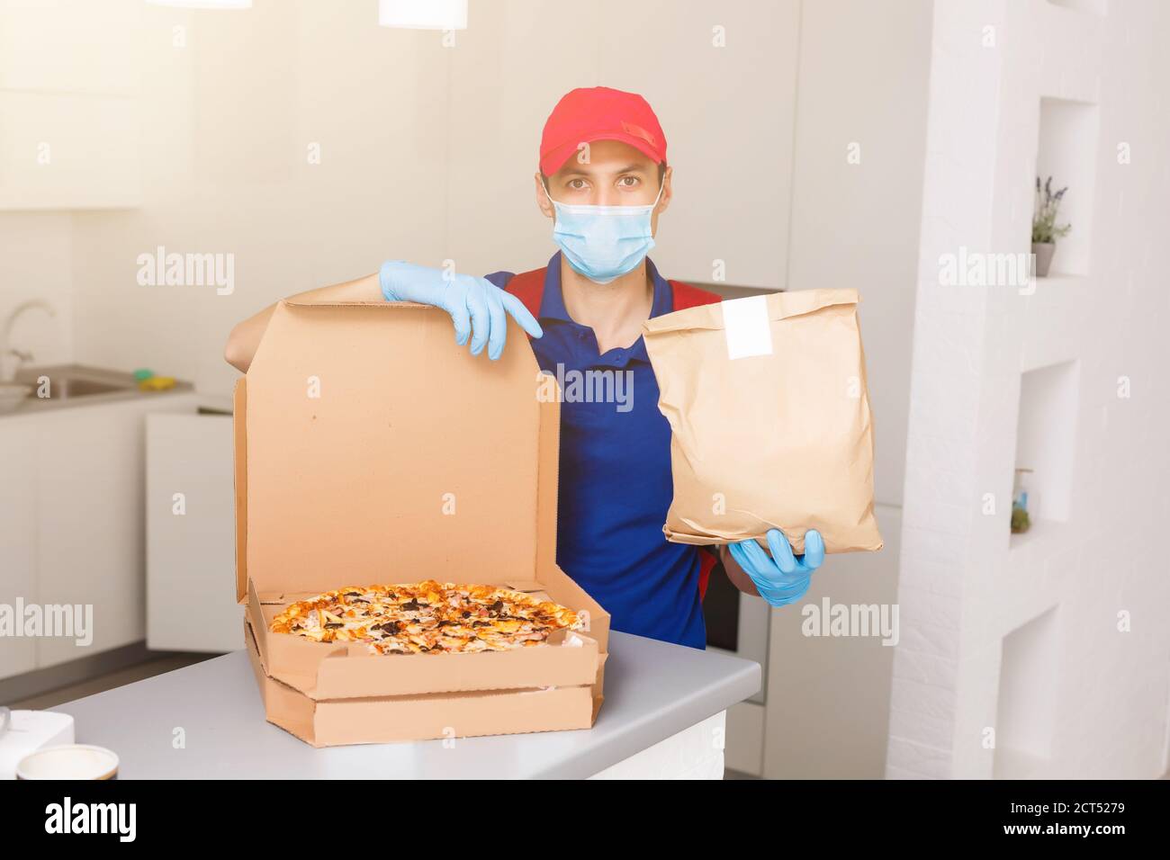 Delivery man employee in red cap t-shirt uniform mask gloves give food order pizza boxes isolated on yellow background studio. Service quarantine Stock Photo