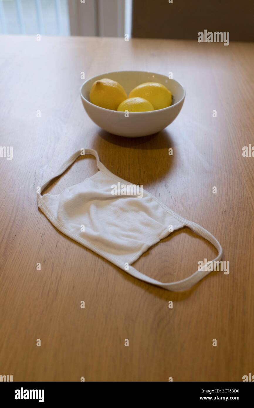 a reusable white cotton facial mask to combat Covid 19 lies on a domestic kitchen table next to a white china bowl with three fresh lemons in it Stock Photo